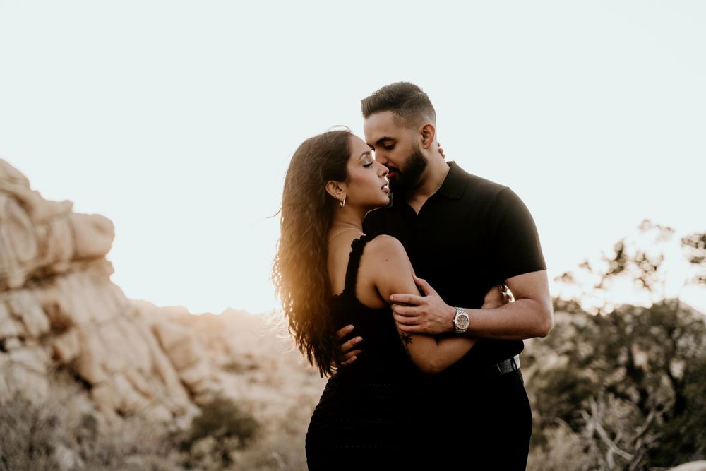 A romantic couple dressed in black embraces during a desert sunset photoshoot among rocky terrain.