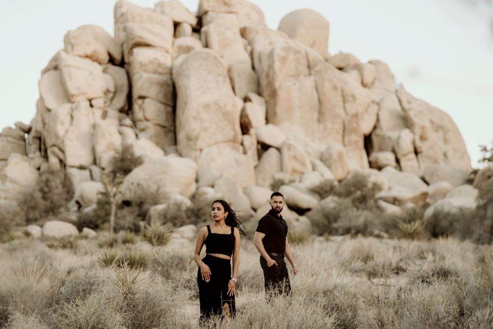 Two people stand in desert brush against towering rock formations at Joshua Tree National Park.