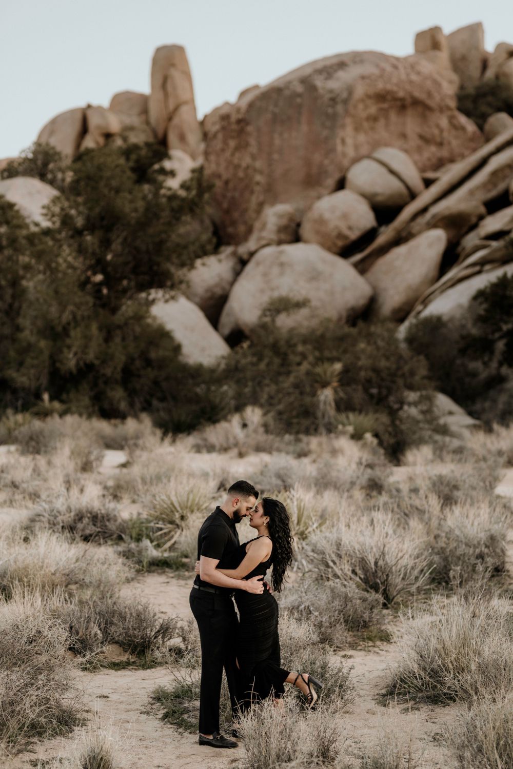 Couple embraces in desert landscape with large rock formations at Joshua Tree National Park during sunset.