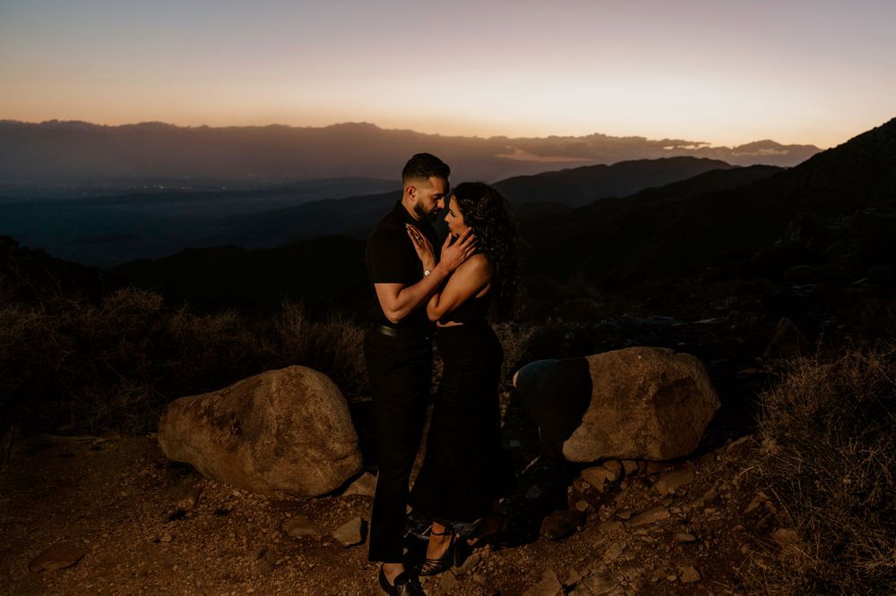 Silhouette figure in black clothing sits on rocky outcrop against mountainous sunset landscape backdrop.