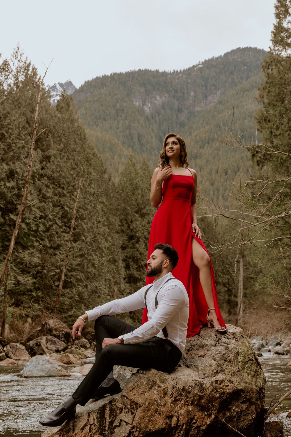 Two people pose dramatically in formalwear on rocks by a mountain stream with evergreen forest in the background.