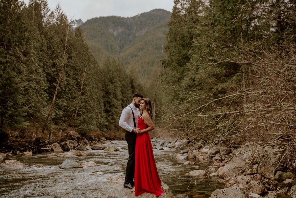 A couple embraces along a rushing mountain river surrounded by evergreen forest in moody atmospheric lighting.