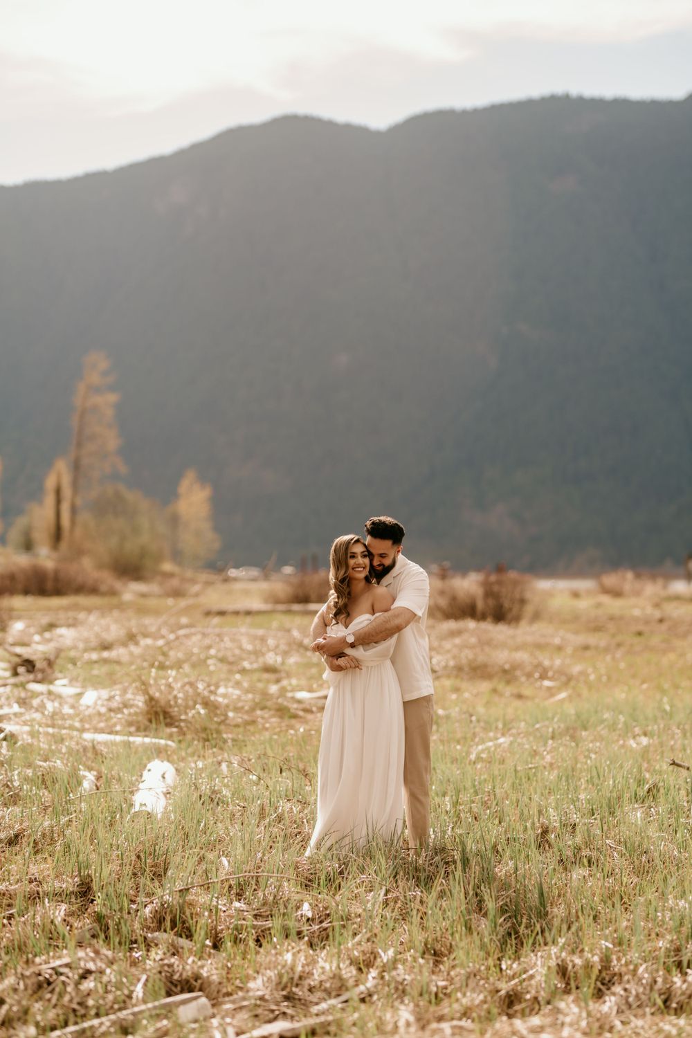 A romantic couple embraces in a flowing white dress against a mountain landscape at sunset in a wildflower meadow.