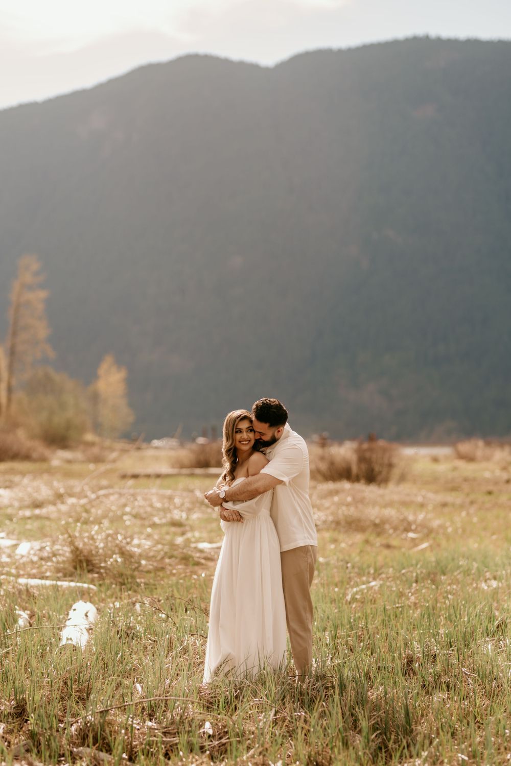 A couple shares an intimate embrace in a golden meadow with misty mountains in the background at sunset.