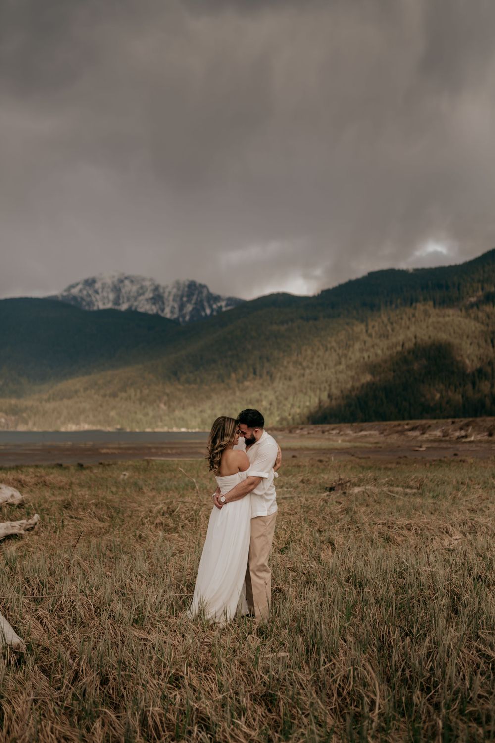A romantic couple embraces in a field with dramatic mountain landscape and moody clouds in the background.