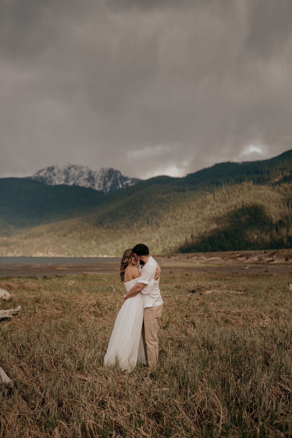 A couple embraces in a field with dramatic mountain peaks and moody skies in the background.