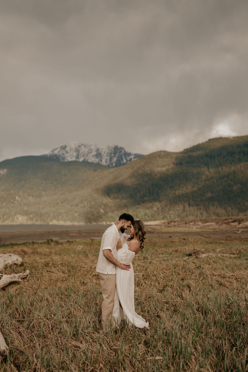 A romantic pair embraces in an outdoor field with dramatic mountains and moody skies in the background.