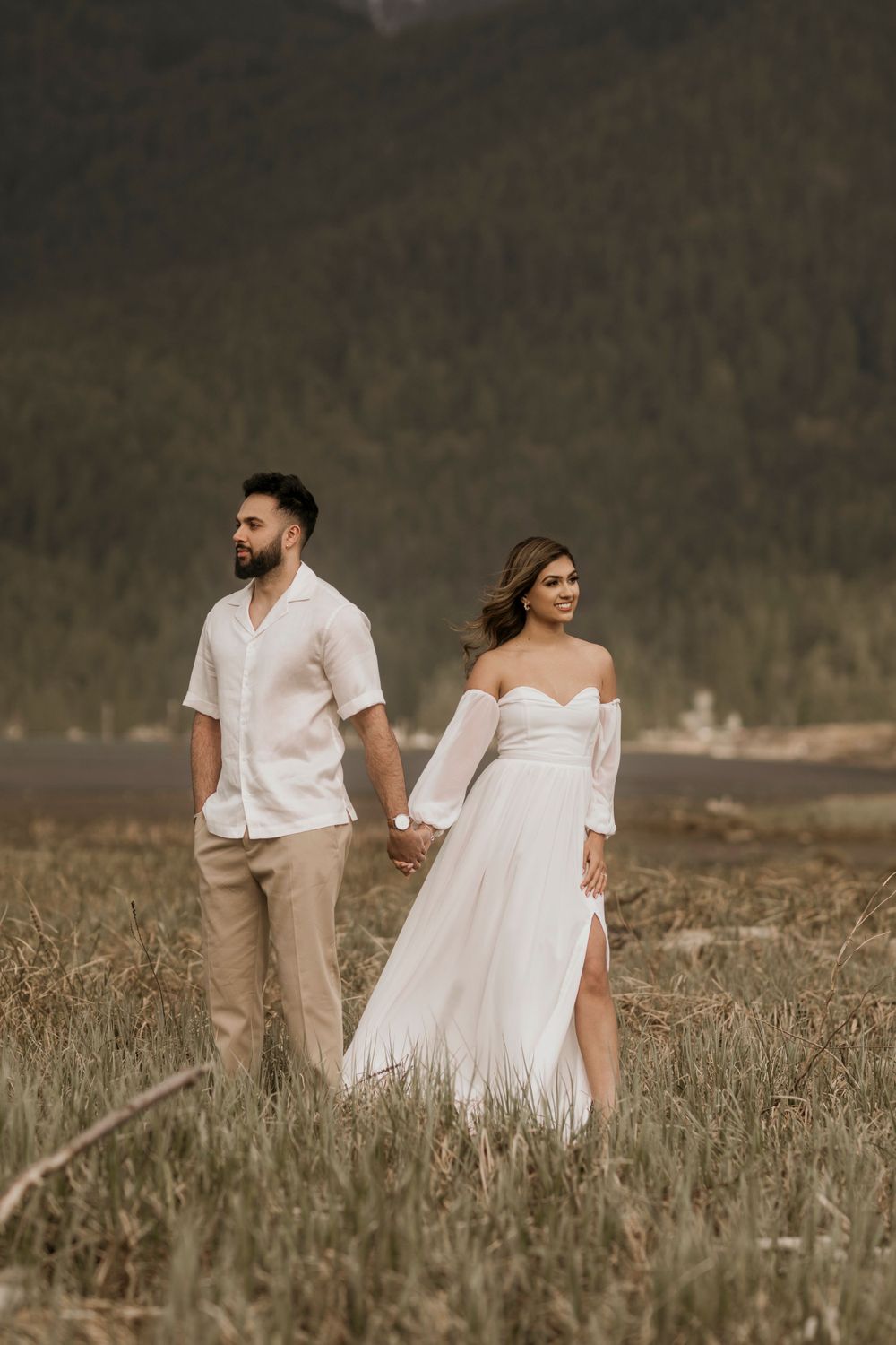 A couple in white attire hold hands while standing in a meadow with mountains in the background at golden hour.