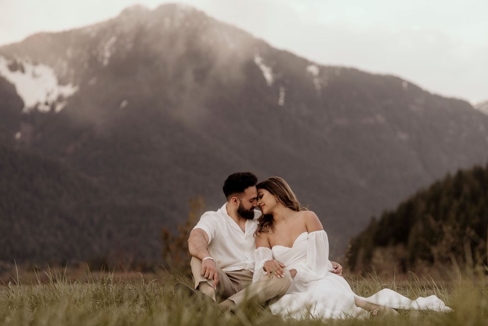 A romantic couple sits in a mountainous meadow with snow-capped peaks in the background at sunset.