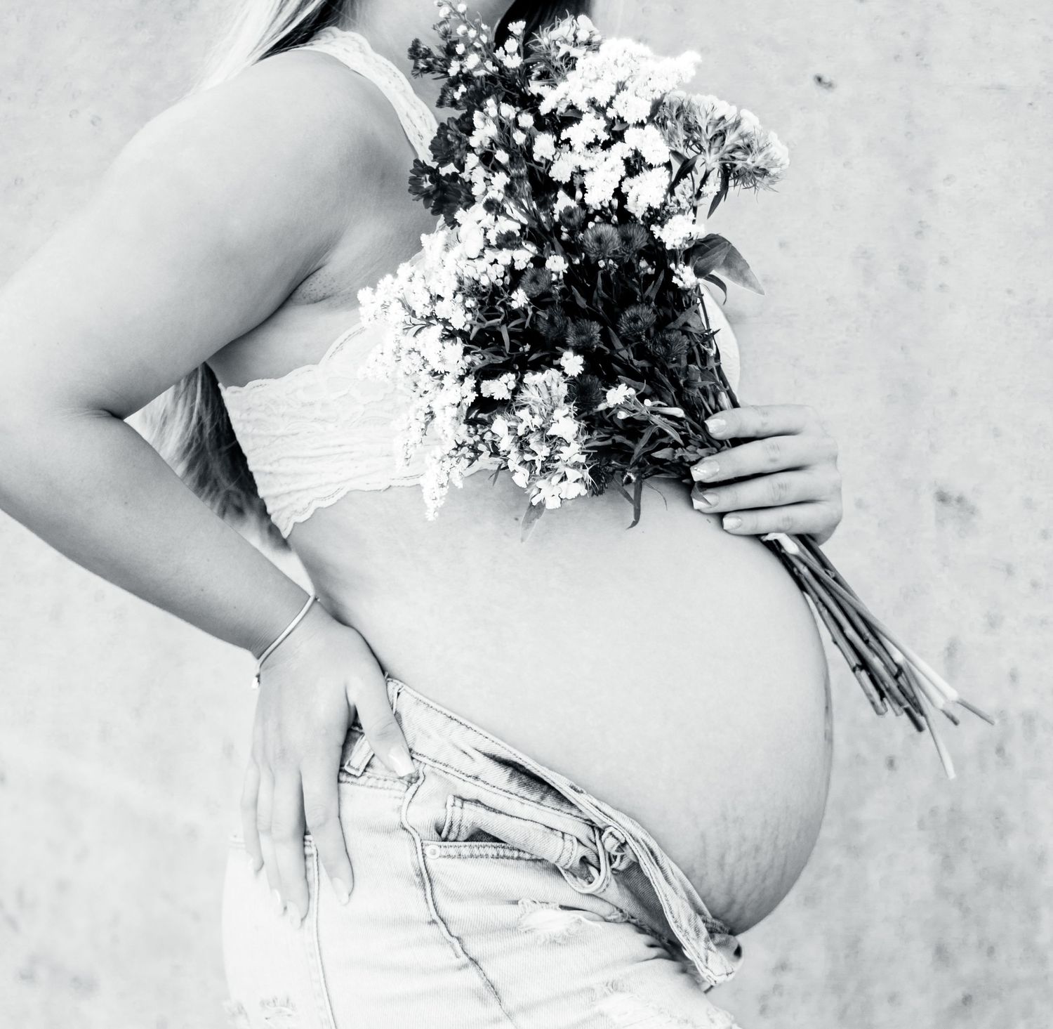 Black and white photo of pregnant person in white dress holding small bouquet of white flowers against light wall.