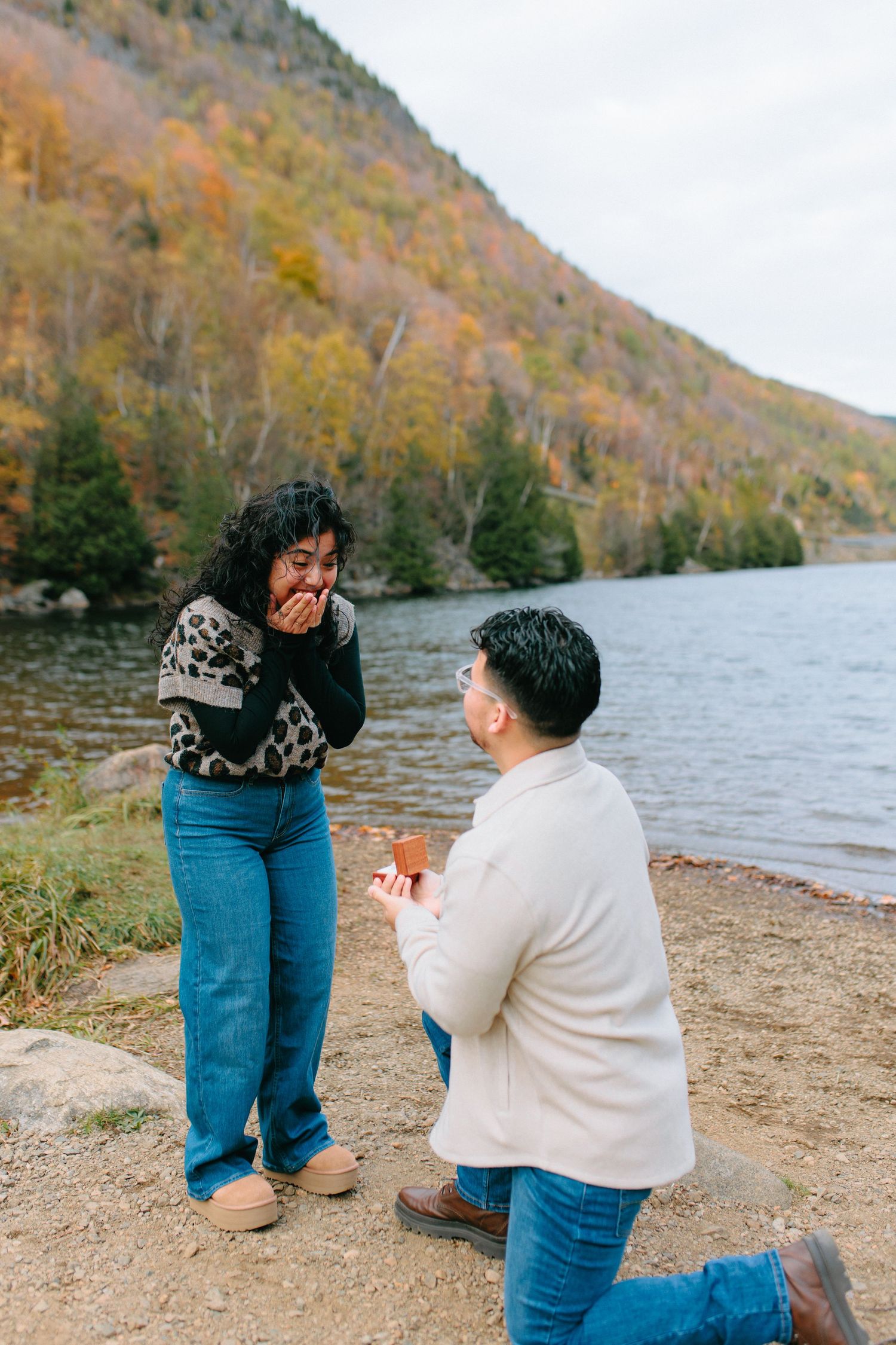 A romantic proposal moment unfolds on a lakeside beach with autumn mountains in the background.