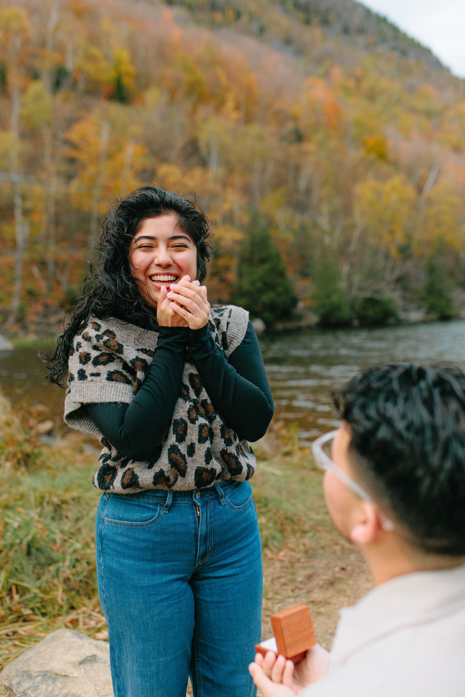 A couple shares a joyful candid moment outdoors during autumn with fall foliage in the background.