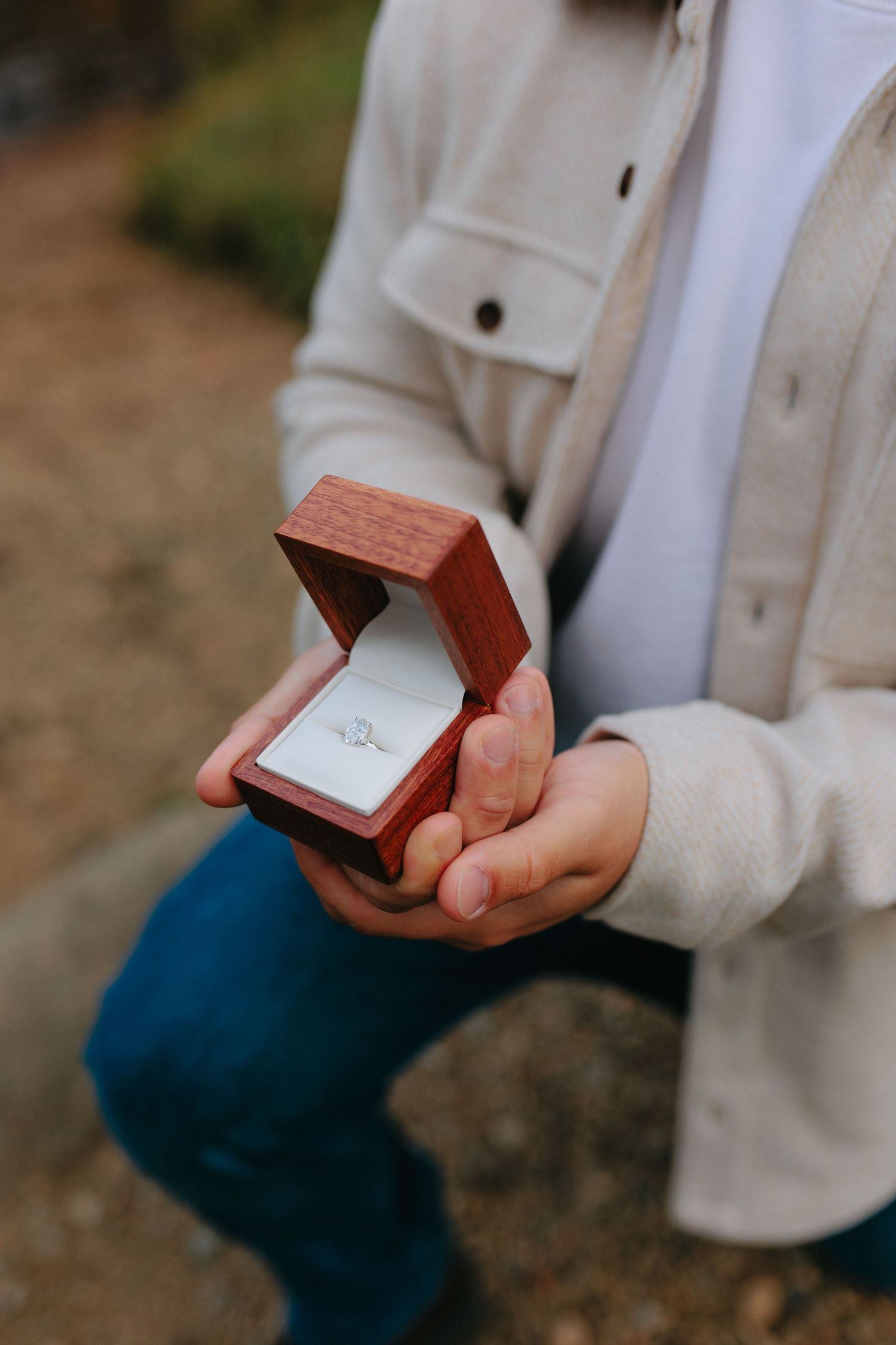 A person wearing a cream colored sweater holds a small wooden ring box in their hands against a blurred background.