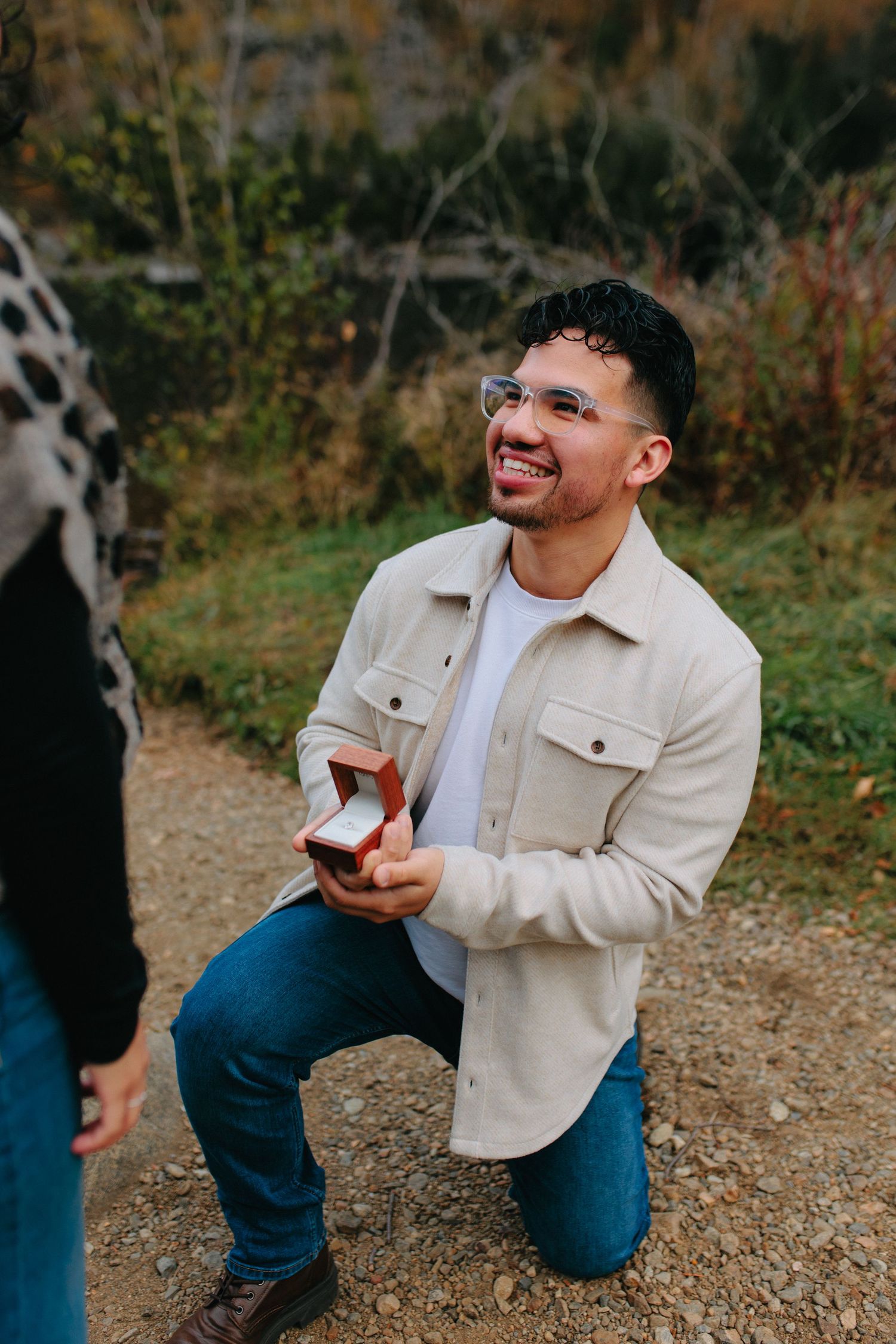 A person in a beige jacket and jeans kneels on a gravel path while holding a wine glass in an outdoor setting.