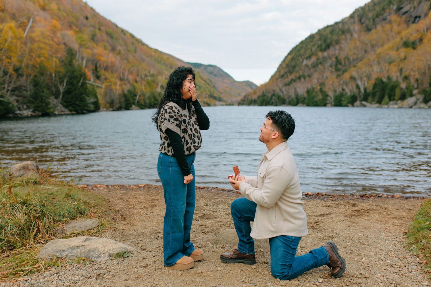 A romantic proposal unfolds on a scenic lakeshore with autumn mountains in the background during golden hour.