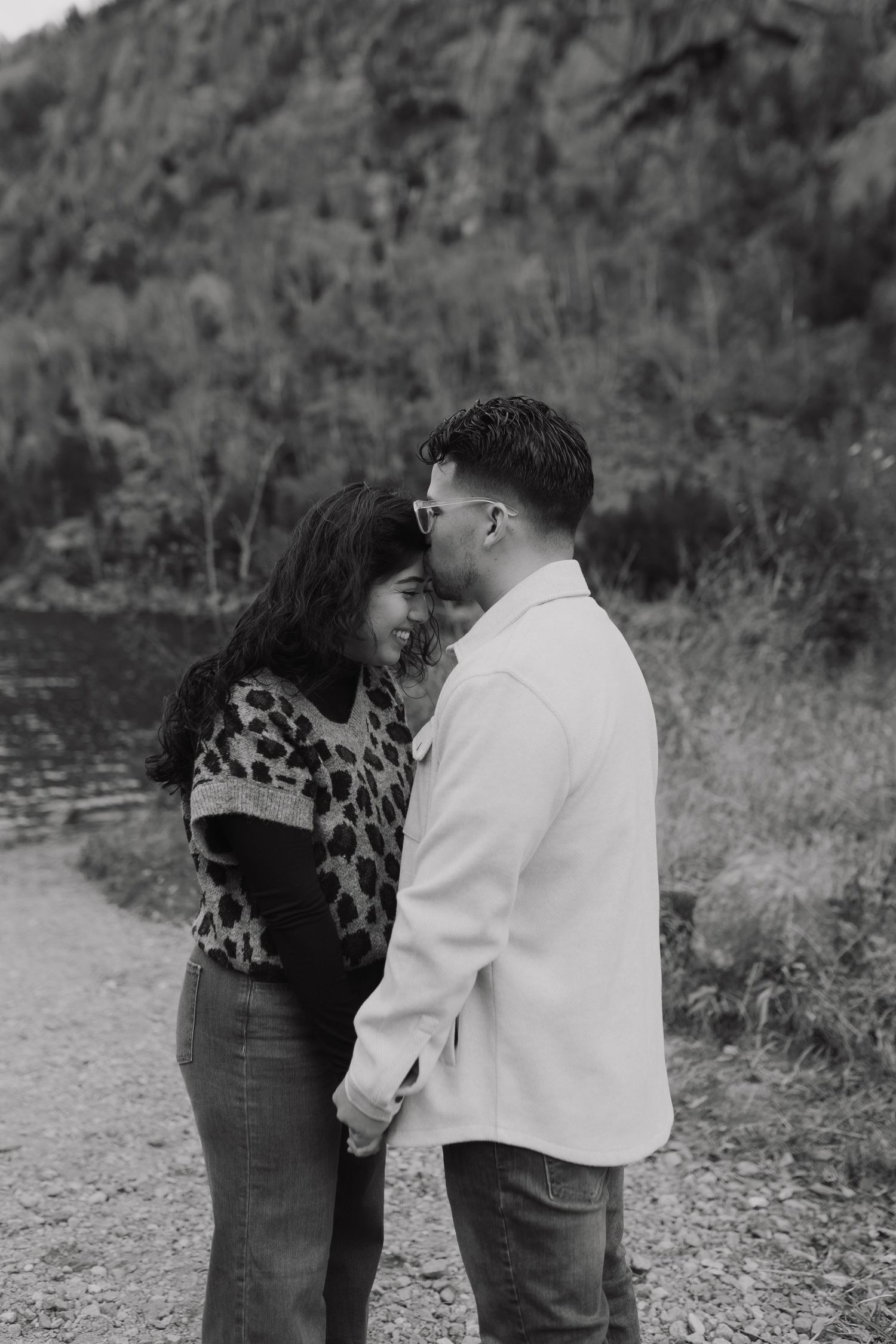A romantic black and white portrait of a couple sharing an intimate moment by a lake with mountains in the background.