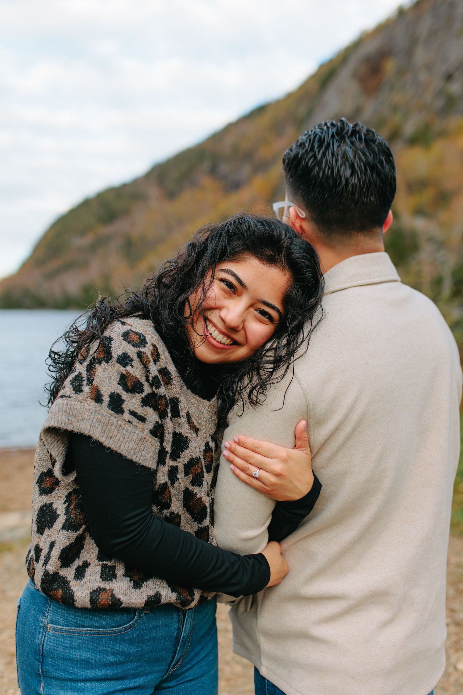 A couple embraces and laughs together against a scenic autumn landscape with water in the background.