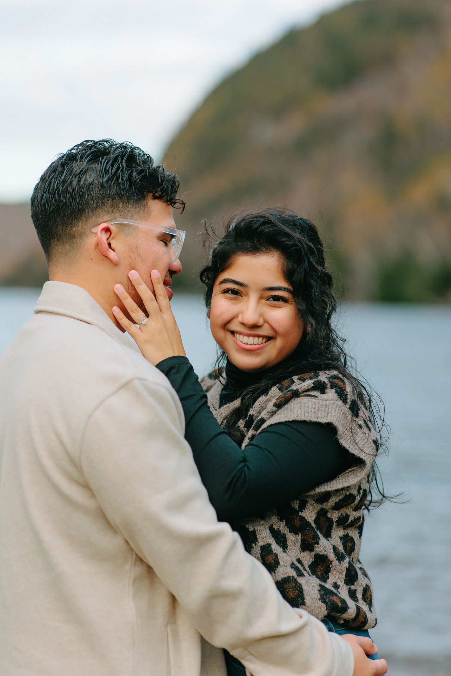 Two people embrace lovingly against a backdrop of mountains and lake waters during autumn.