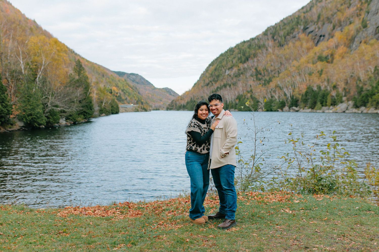 A couple stands together by a scenic mountain lake surrounded by autumn foliage on a cloudy day.