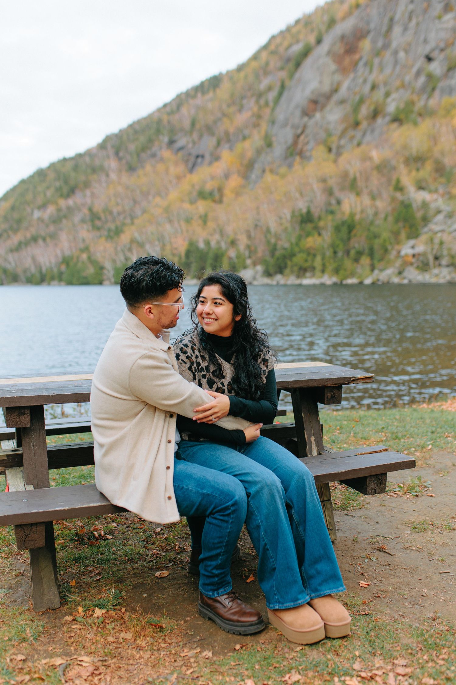 A couple sits together on a wooden bench overlooking a serene mountain lake.