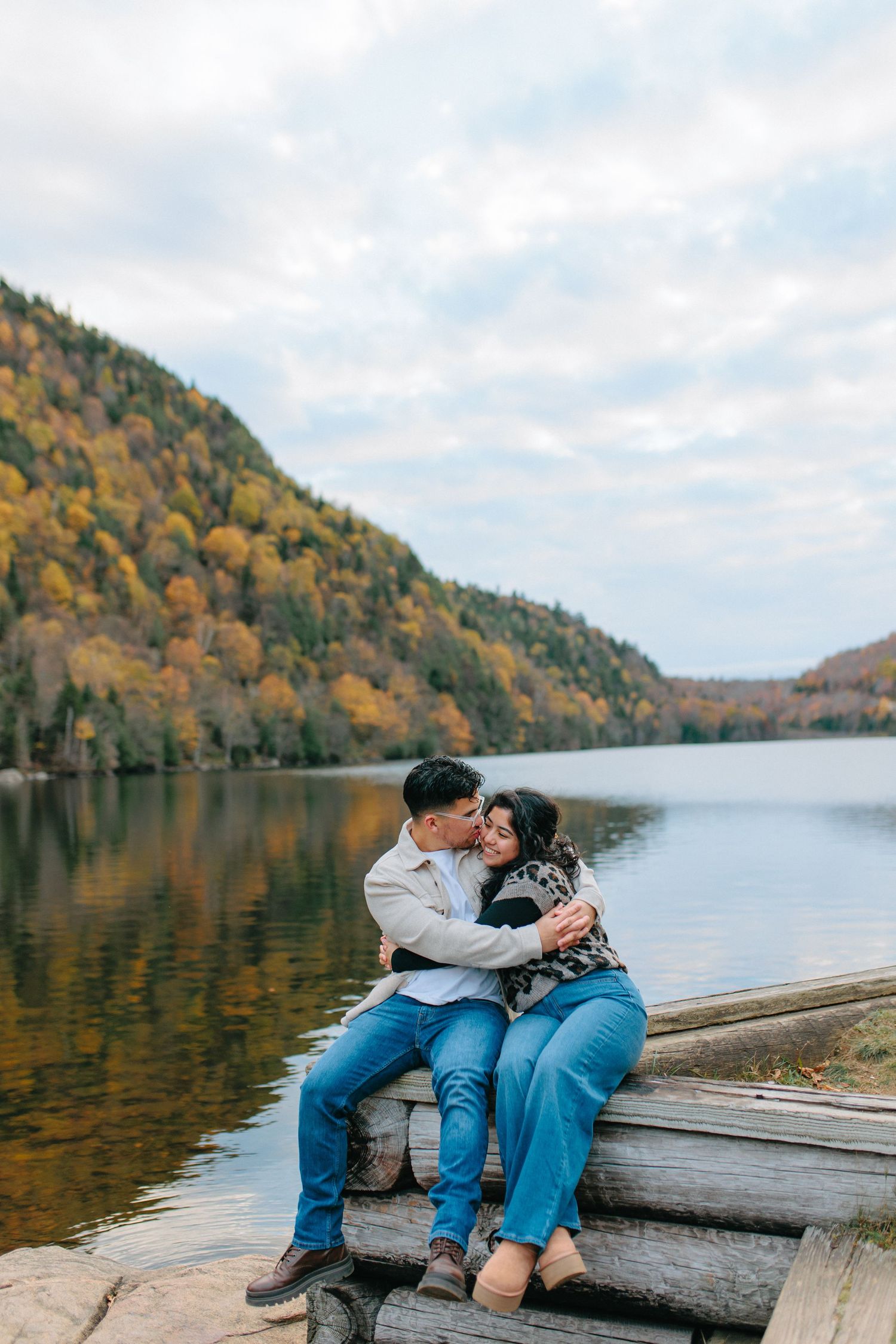 A couple sits close together on a wooden dock overlooking a serene lake surrounded by fall-colored mountains.