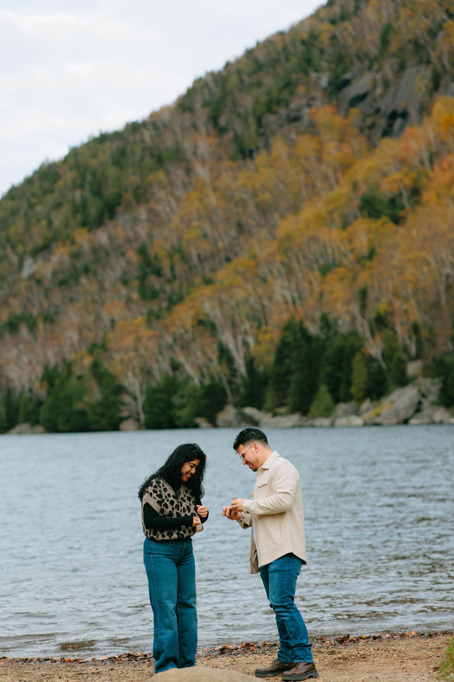 Two people stand on a lakeside beach looking at their phones with autumn foliage covered mountains in background.
