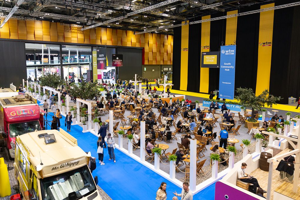 Aerial view of a busy indoor event space with food trucks, vendor booths and attendees on a blue floor with yellow walls.