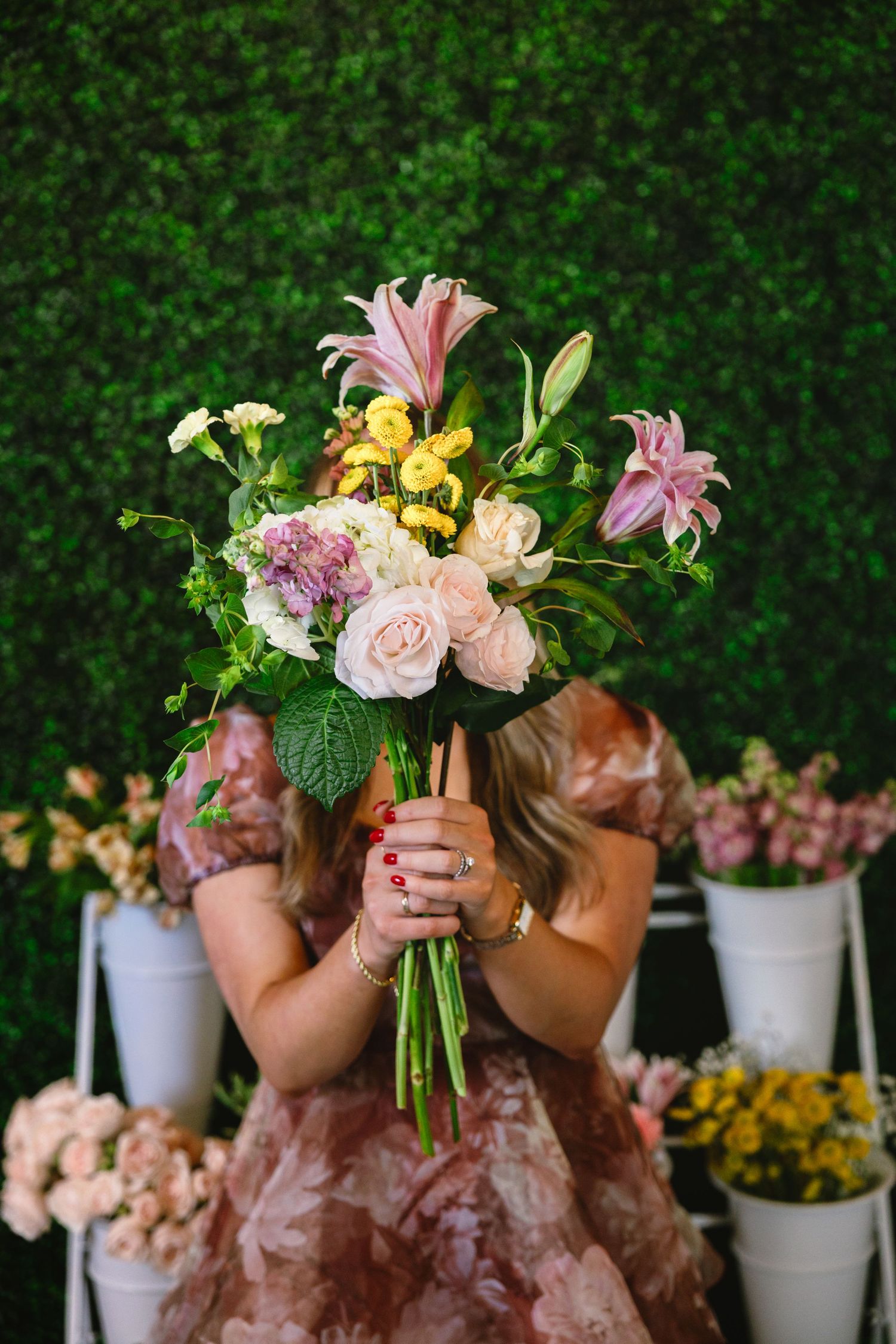 A florist arranges fresh pink, yellow and white flowers into a bouquet against a green hedge background.