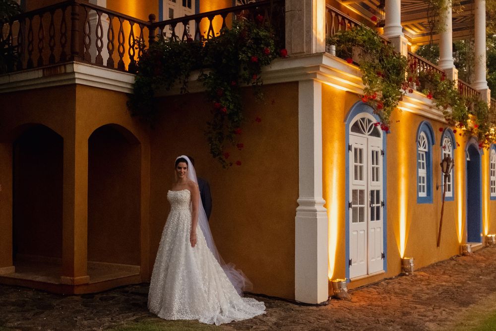 A bridal figure stands in a warmly lit colonial-style courtyard with arched columns and stone walkways at night.