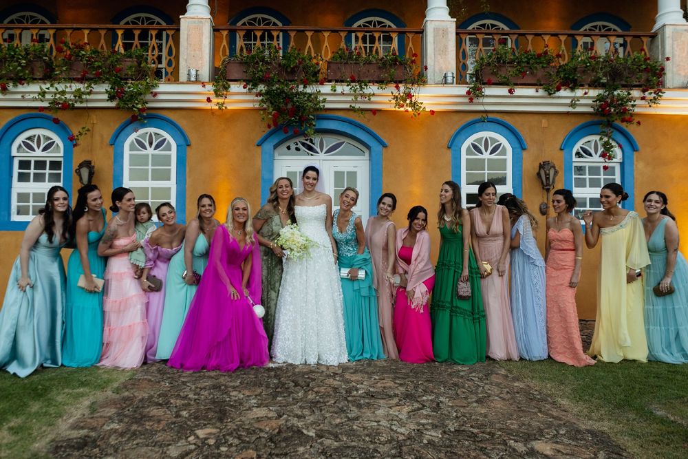 Large wedding party in colorful formal attire poses in front of yellow building with white arched windows.