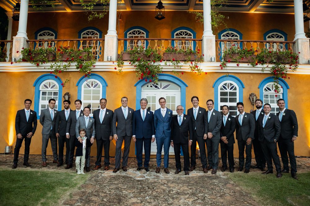 Wedding party in formal black suits and dresses lined up against yellow building with white windows.