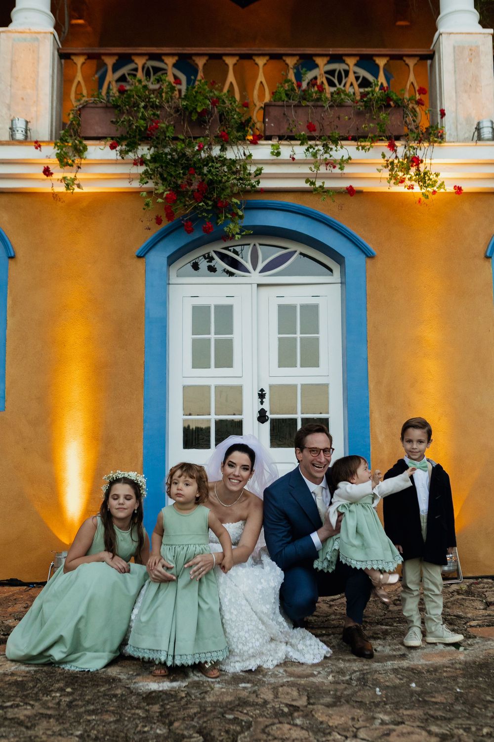 Wedding group photo with children in mint green dresses against yellow building with blue door.