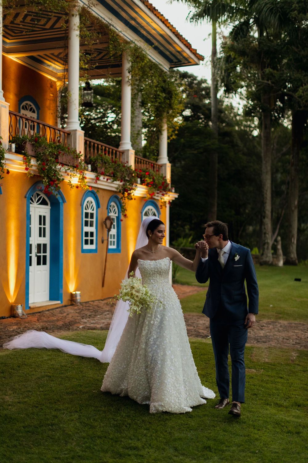 Wedding couple walks together outside a historic yellow colonial building with columns at sunset.