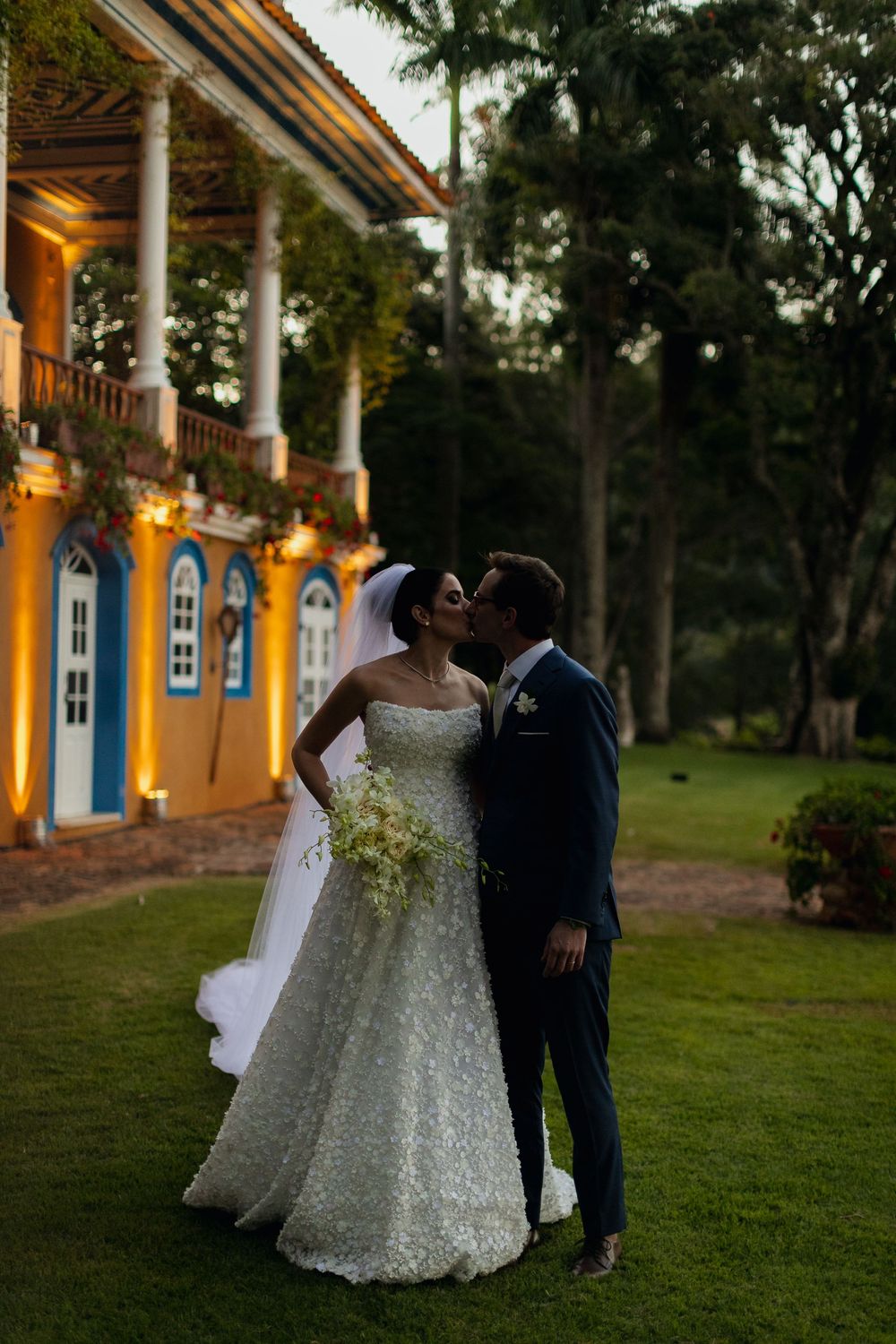 A romantic wedding couple shares intimate moments on a lawn outside a colonial-style building with illuminated pillars at dusk.