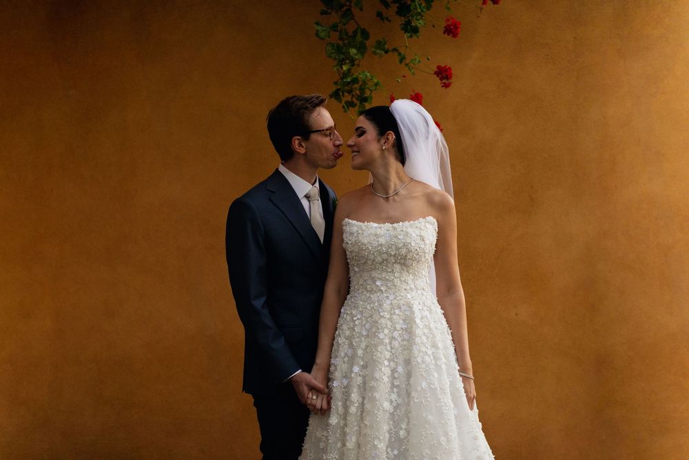 A couple in formal wedding attire shares an intimate moment against a warm brown textured wall.