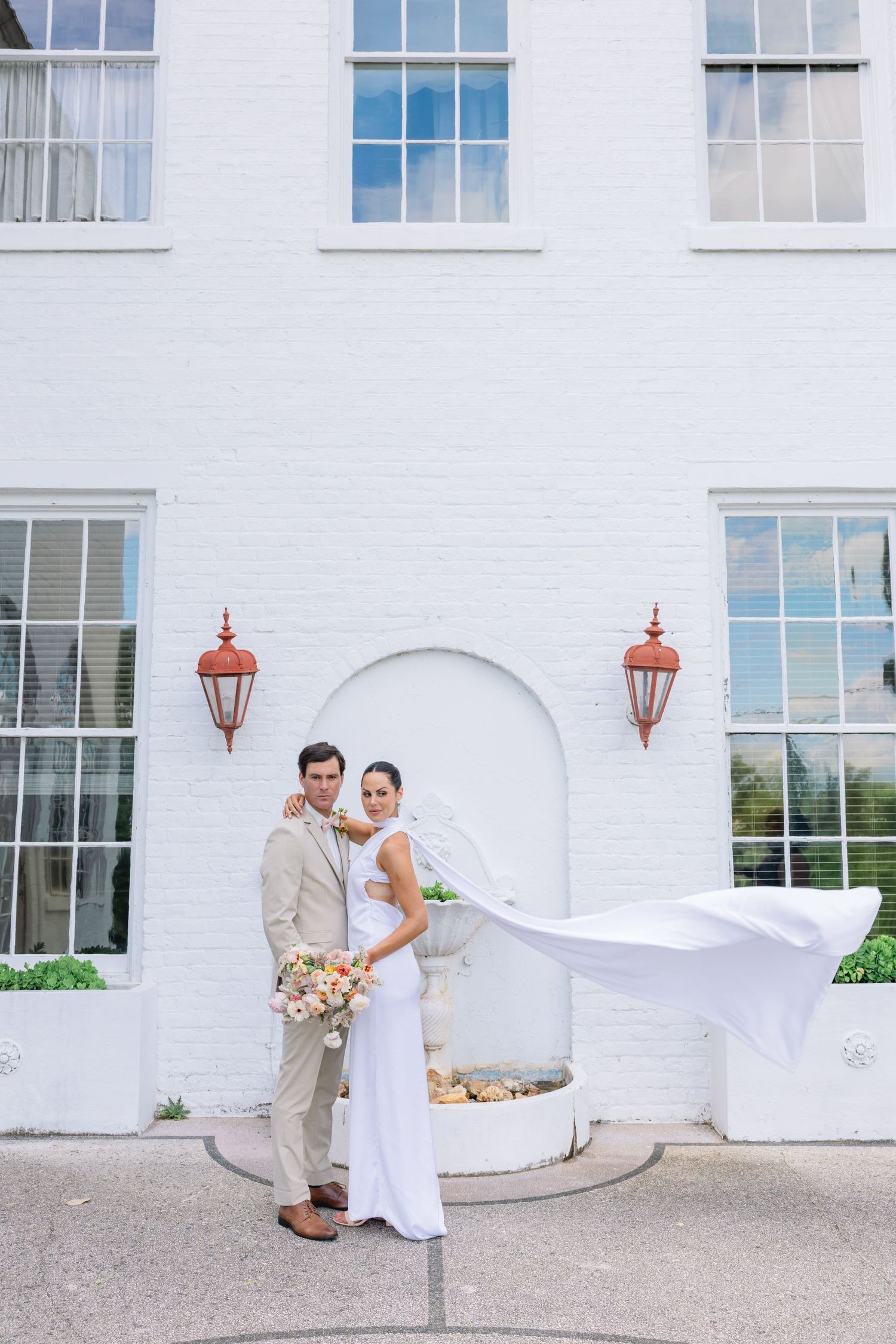 A couple in wedding attire poses against a white building with arched windows and vintage lanterns.