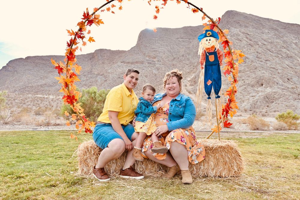 A happy family smiles together on hay bales under a colorful autumn leaf arch.