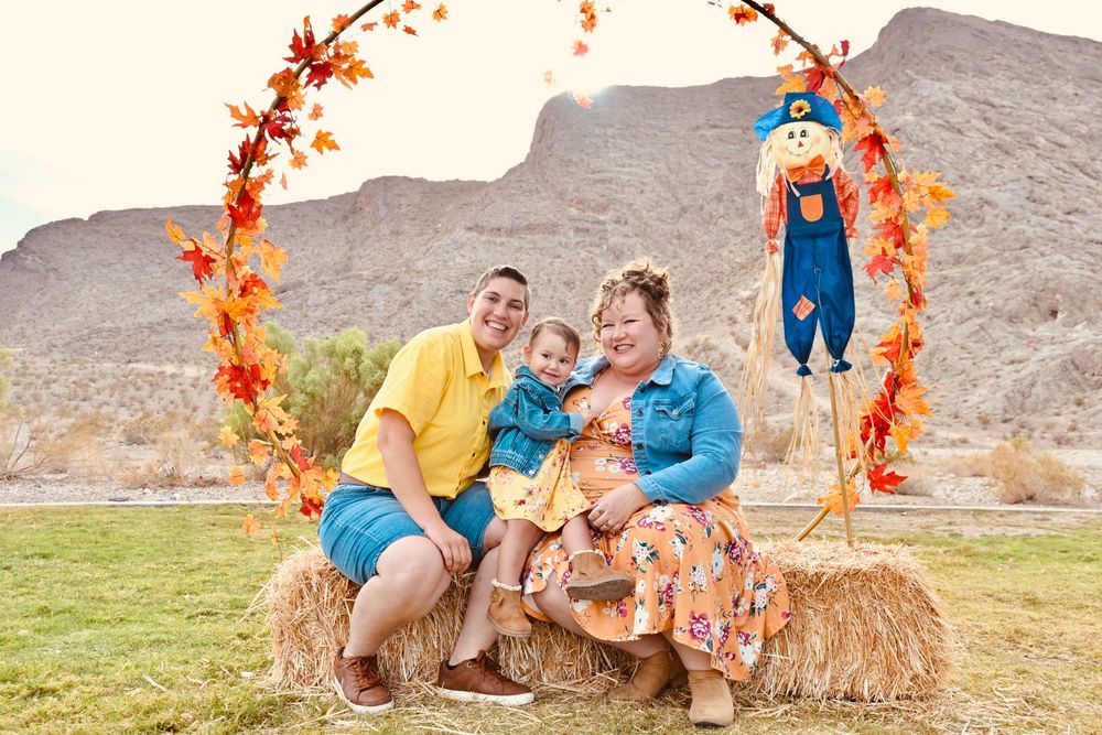 A cheerful family sits on hay bales under an autumn leaf arch with a scarecrow.