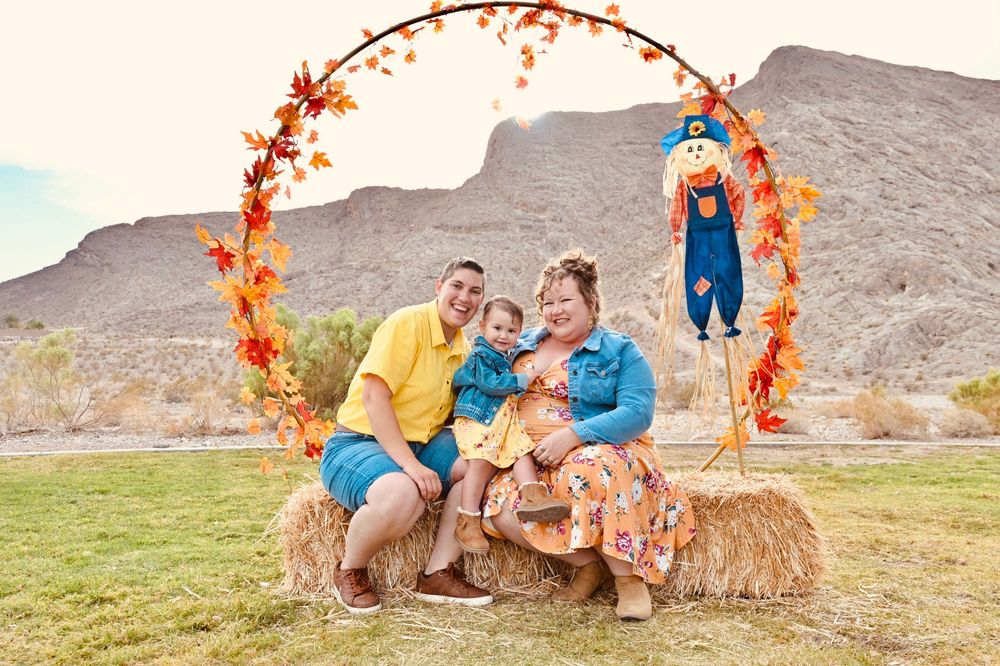 Family of three poses on hay bale under autumn leaf arch with scarecrow in desert landscape.