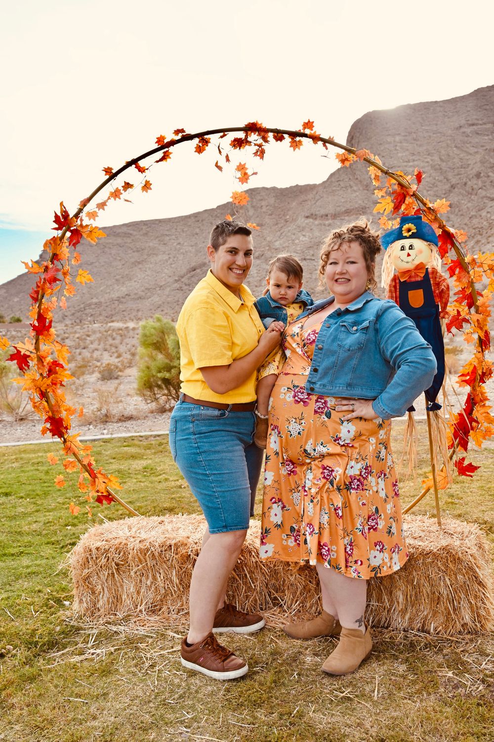Family of three posing on hay bale under autumn leaf arch with scarecrow in desert landscape.