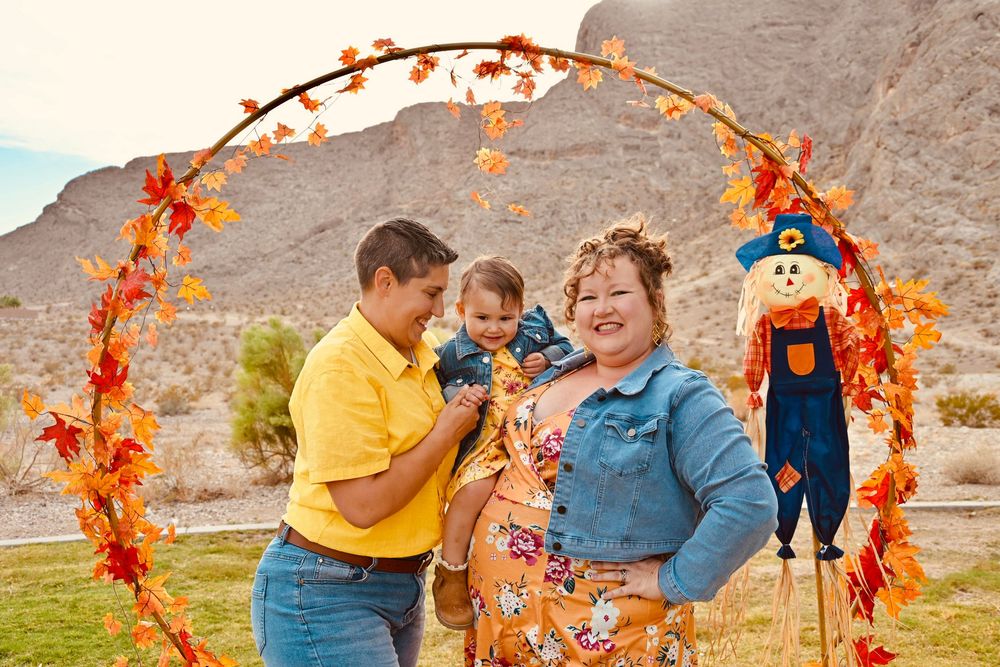 Smiling family of three standing under decorative autumn arch with scarecrow in desert setting.
