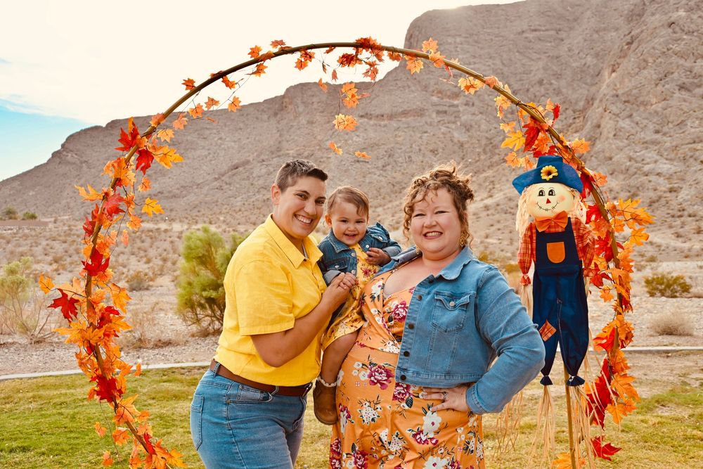 Family of three smiling together under orange autumn leaf arch with scarecrow in desert backdrop.
