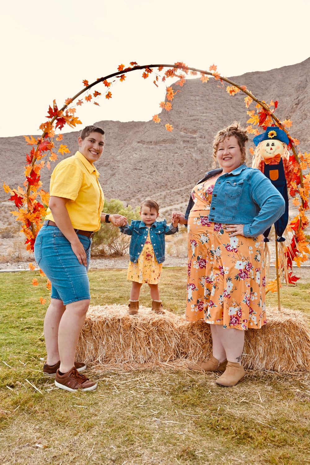 Family posing on hay bales under an autumn leaf arch with scarecrow in a desert landscape.