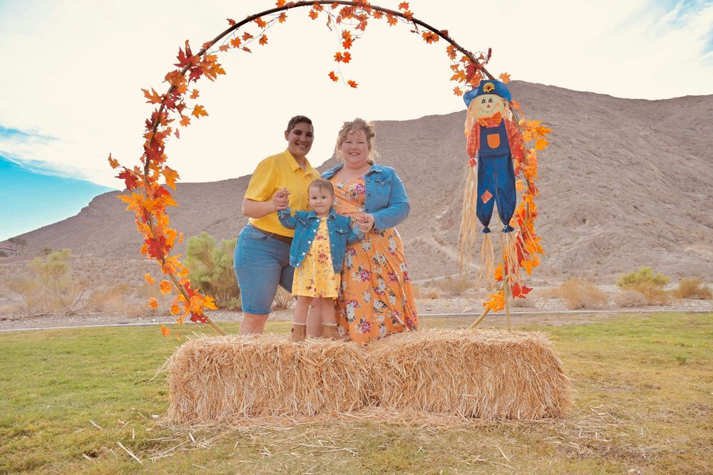 Family of three standing on hay bales under a colorful fall leaf arch in a desert setting.