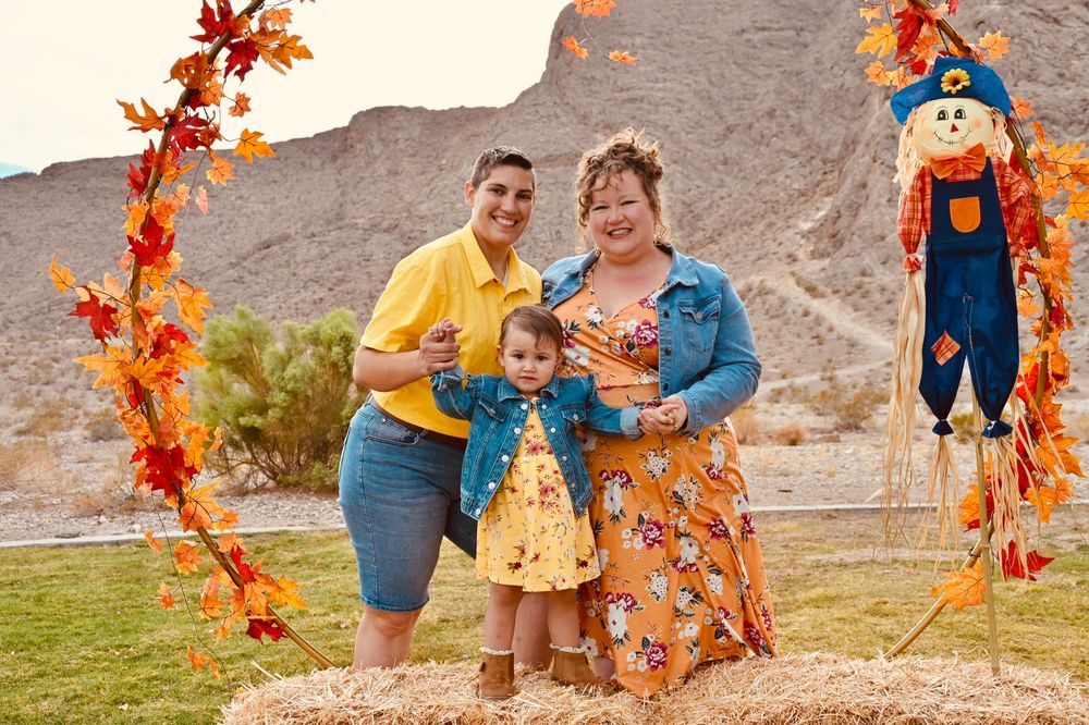Smiling family of three posing beside a scarecrow under an autumn arch on a hay bale.
