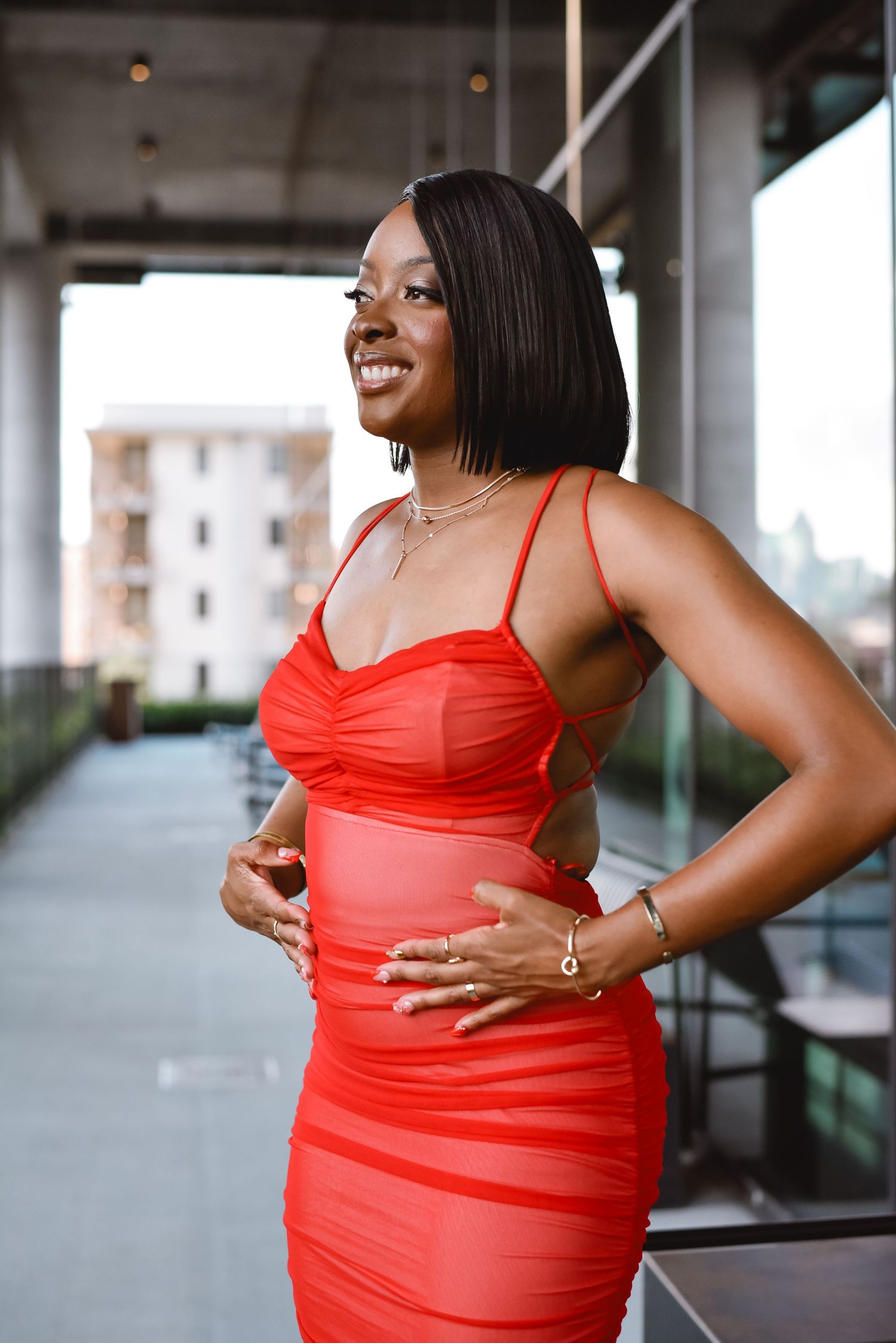 Woman in bright red dress poses confidently in an urban setting with natural lighting and architectural elements.