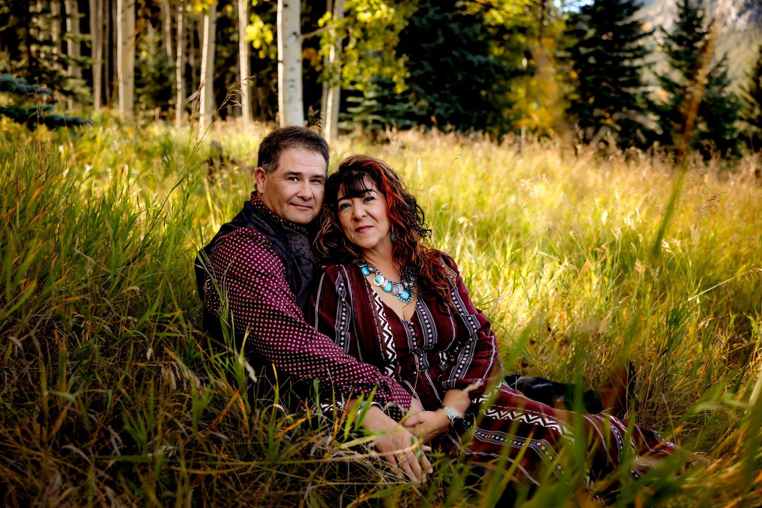 Couple sitting together in a golden autumn meadow surrounded by evergreen trees during sunset.