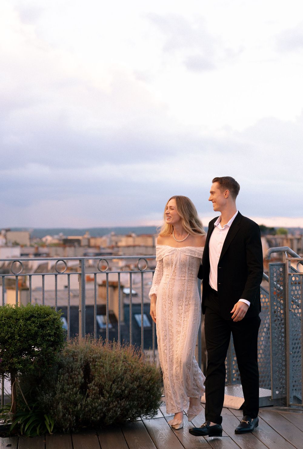 Couple in formal attire sharing romantic moments on a rooftop terrace with city buildings in the background at sunset.