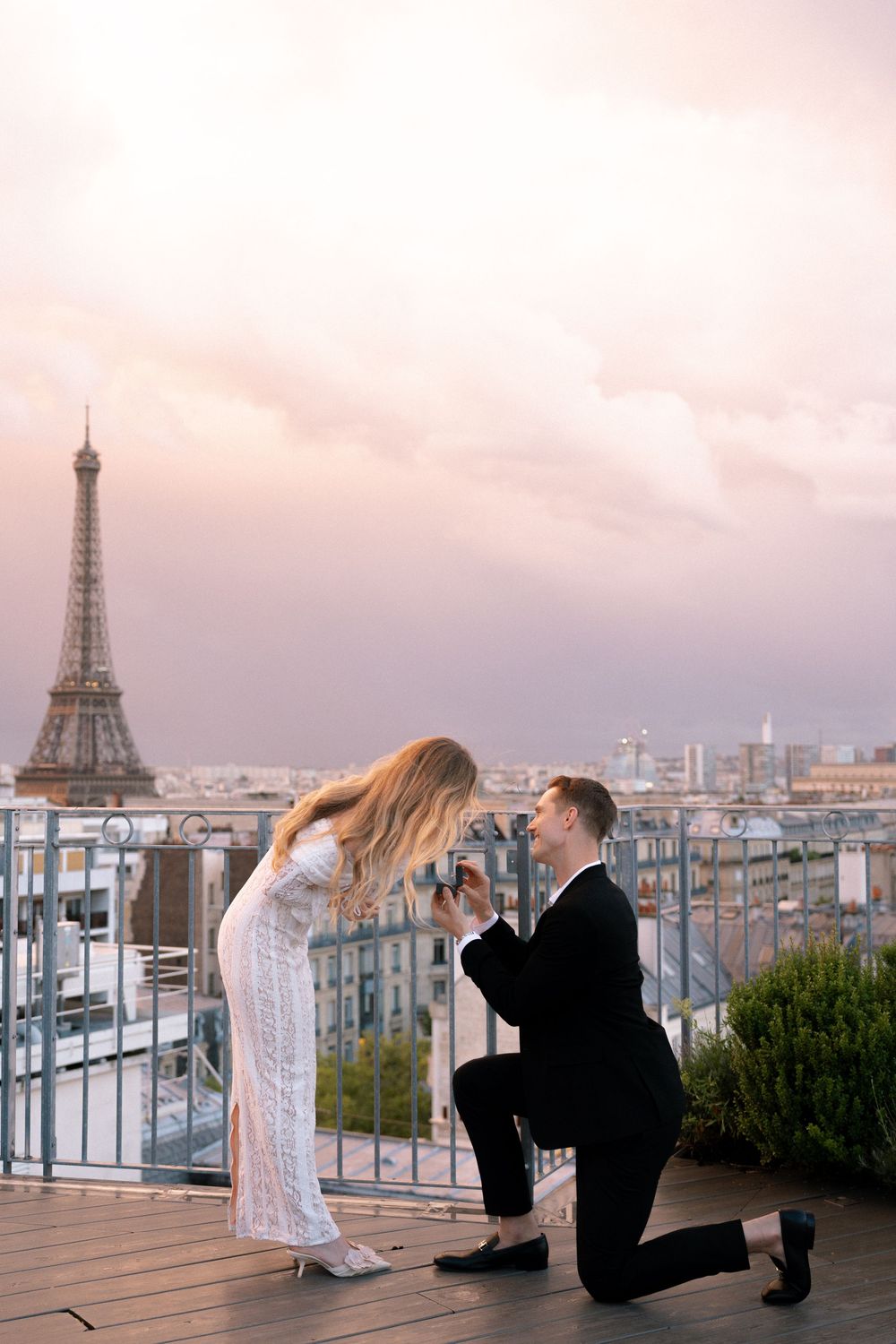 A romantic proposal moment on a rooftop terrace overlooking the Eiffel Tower and Paris cityscape at sunset.