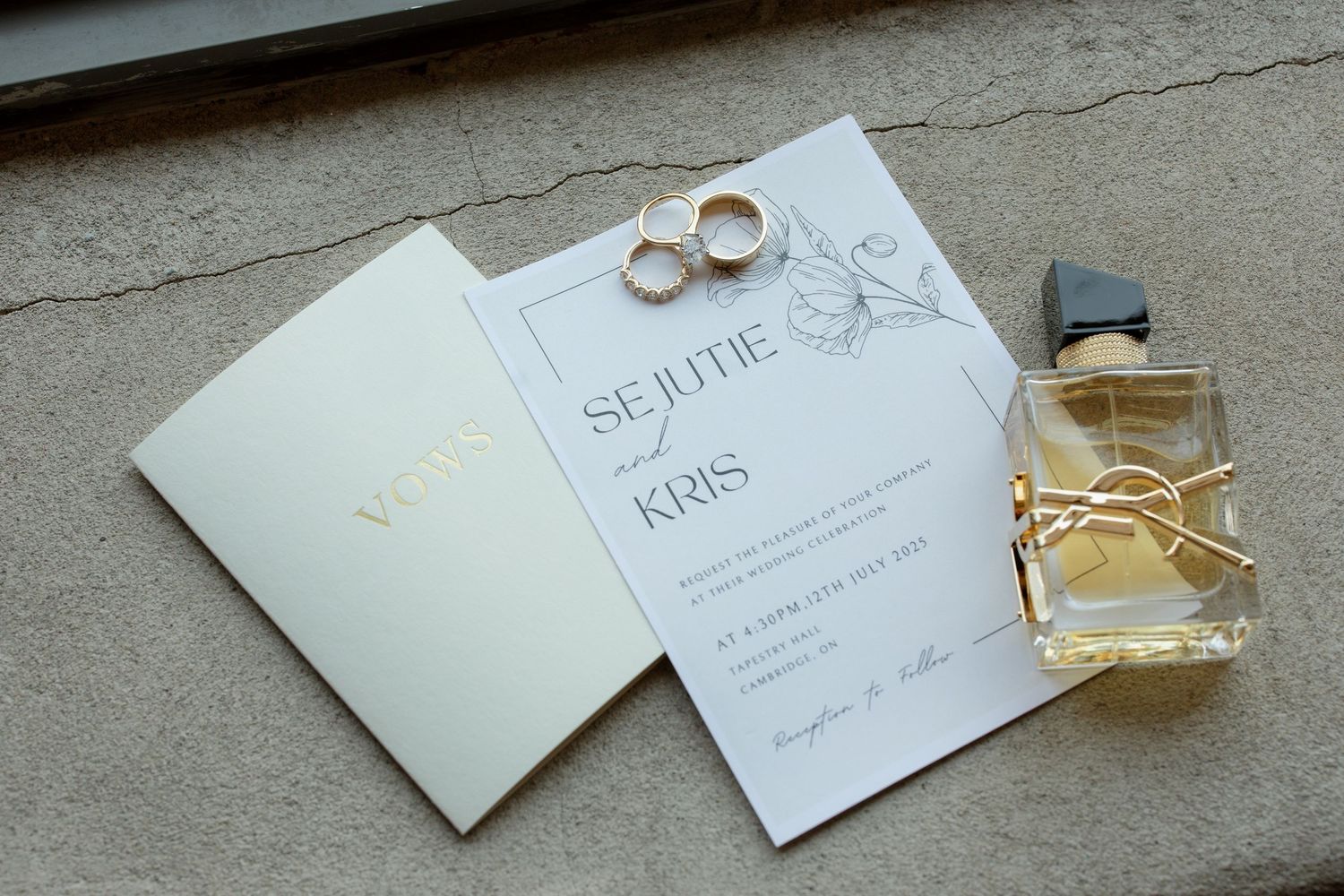 Wedding details including invitation cards, rings, and perfume bottle arranged on a gray surface.